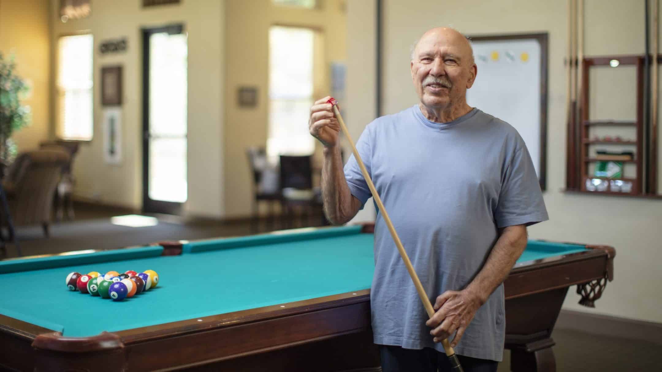 An older man in a light blue shirt smiles while holding a pool cue beside a billiards table with racked balls in a bright, spacious room.