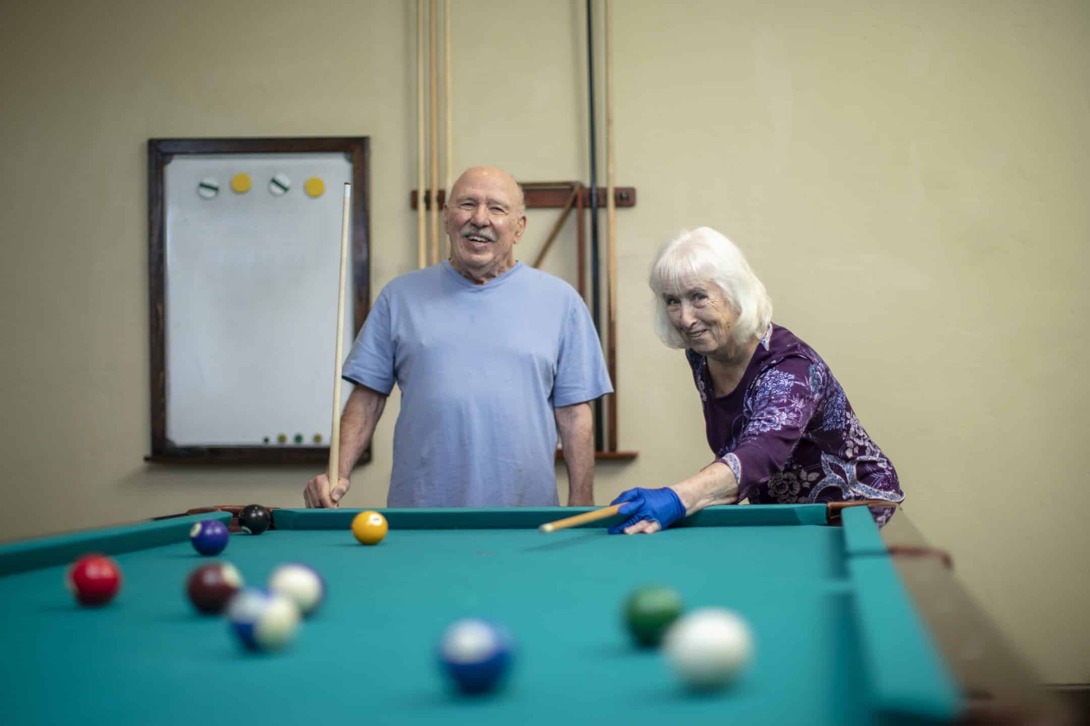 An elderly woman aims her cue at a pool table, preparing to take a shot, while an elderly man stands nearby, smiling. A whiteboard and pool cues are visible on the wall in the background.