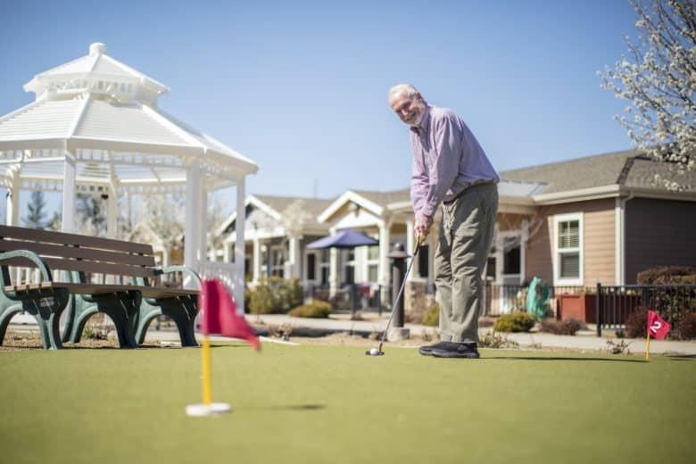 An older man smiles while playing mini-golf on a putting green beside a white gazebo, with benches and houses in the background on a sunny day.