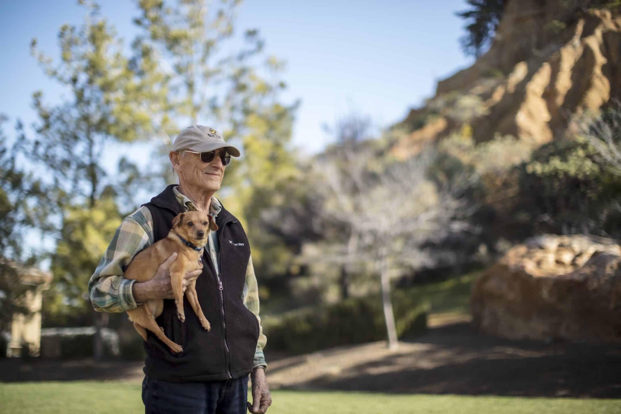 An elderly man wearing sunglasses and a hat stands outdoors, holding a small brown dog. He is in a park-like setting with green grass, trees, and rocky hills in the background on a sunny day.