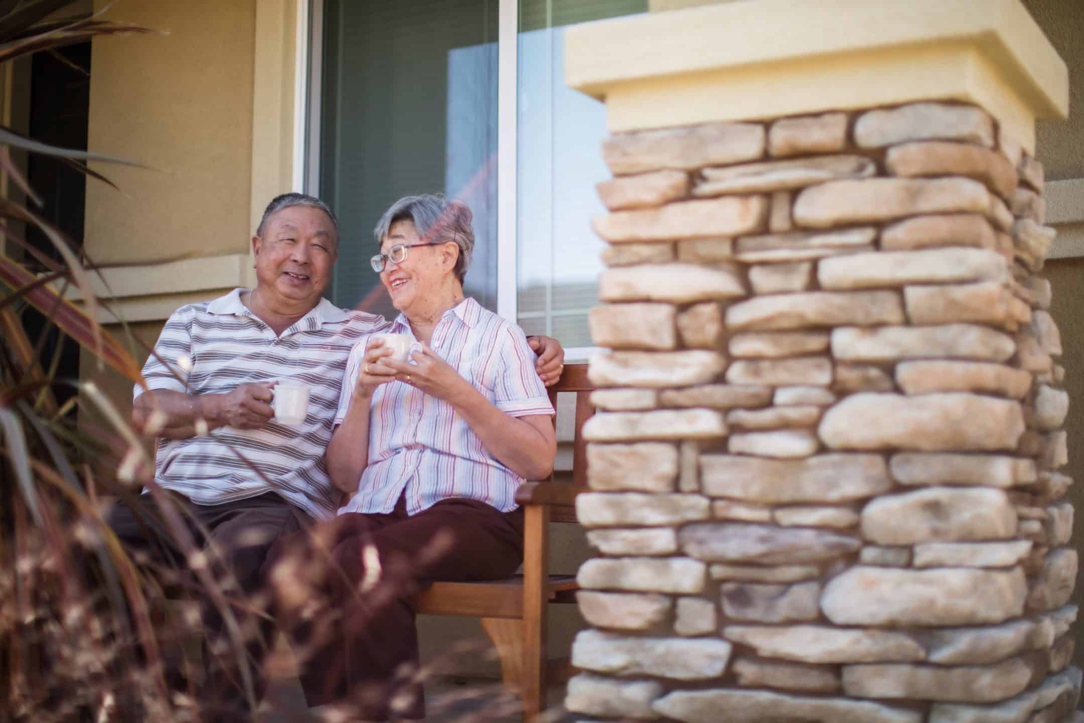 An elderly couple sits on a wooden bench outside, smiling and holding cups, enjoying a relaxed moment together near a stone pillar.