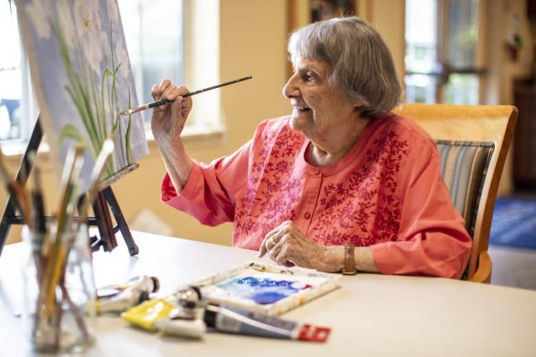 An elderly woman with gray hair, wearing a pink blouse, sits at a table and smiles while painting a flower scene on an easel, surrounded by paint tubes and brushes—a radiant example of Resident Culture in creative aging.