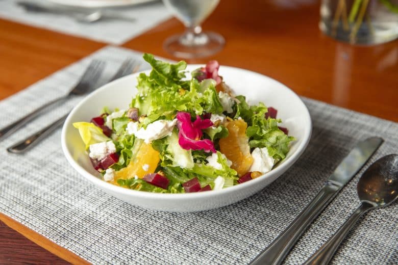 A white bowl filled with a fresh salad of leafy greens, orange slices, beet cubes, white cheese, and edible flower petals, placed on a set dining table with cutlery and a glass of water in the background.