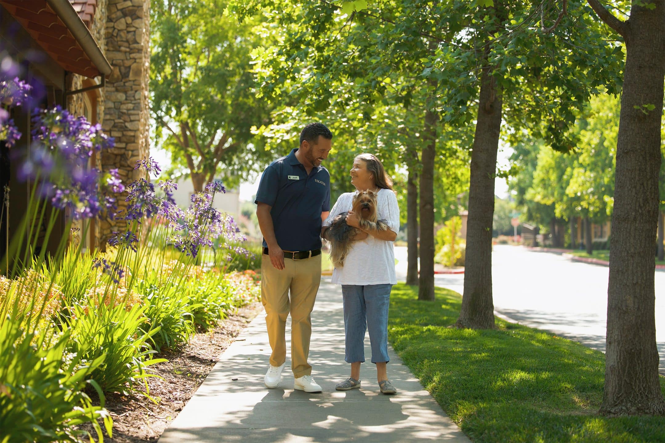A man in a navy shirt and khaki pants walks on a sunny sidewalk with a woman holding a small dog, enjoying assisted living. They smile at each other, surrounded by green trees and purple flowers.