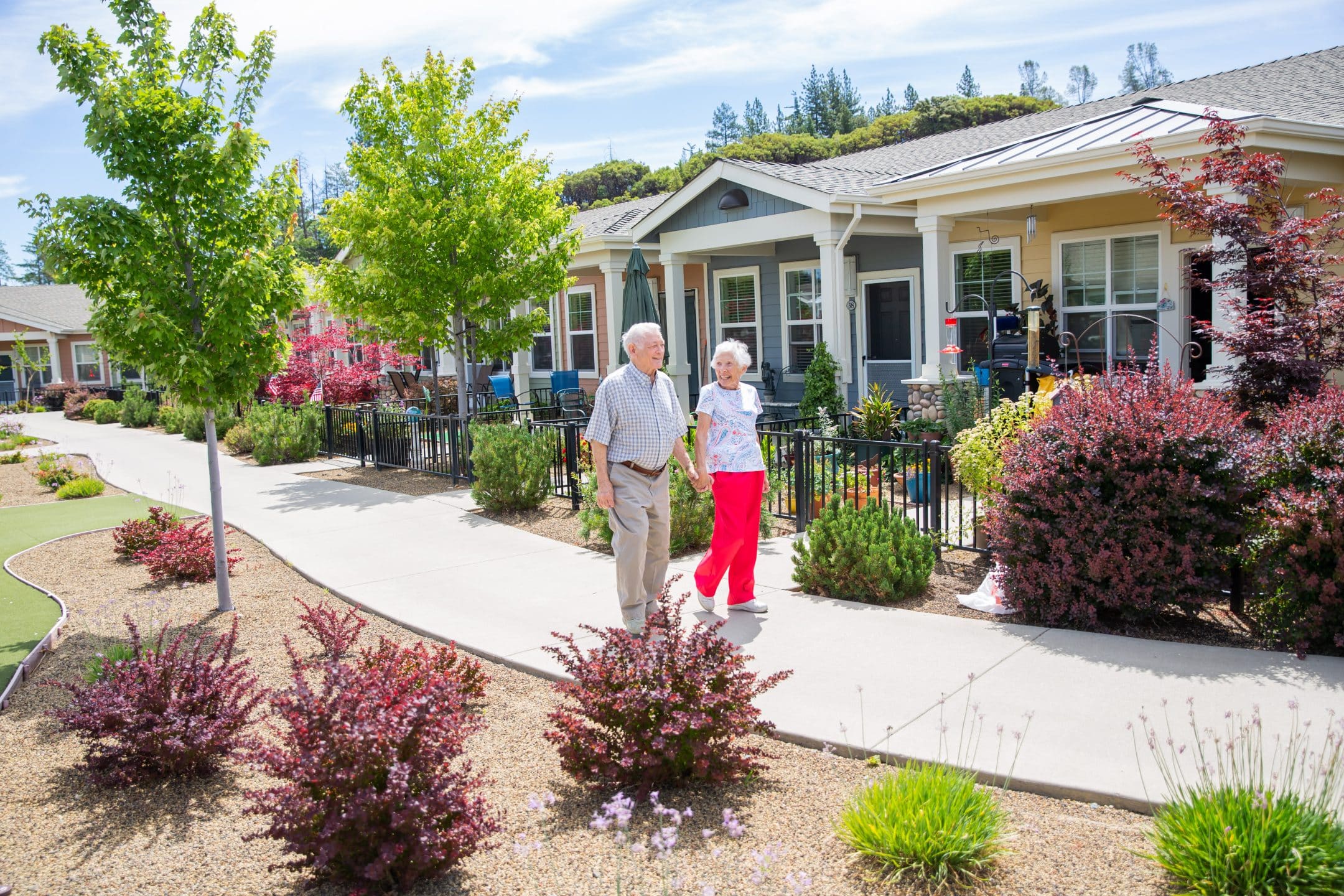 Two seniors walk hand-in-hand on a sunny path through a landscaped garden in front of charming, single-story homes with porches and colorful plants.