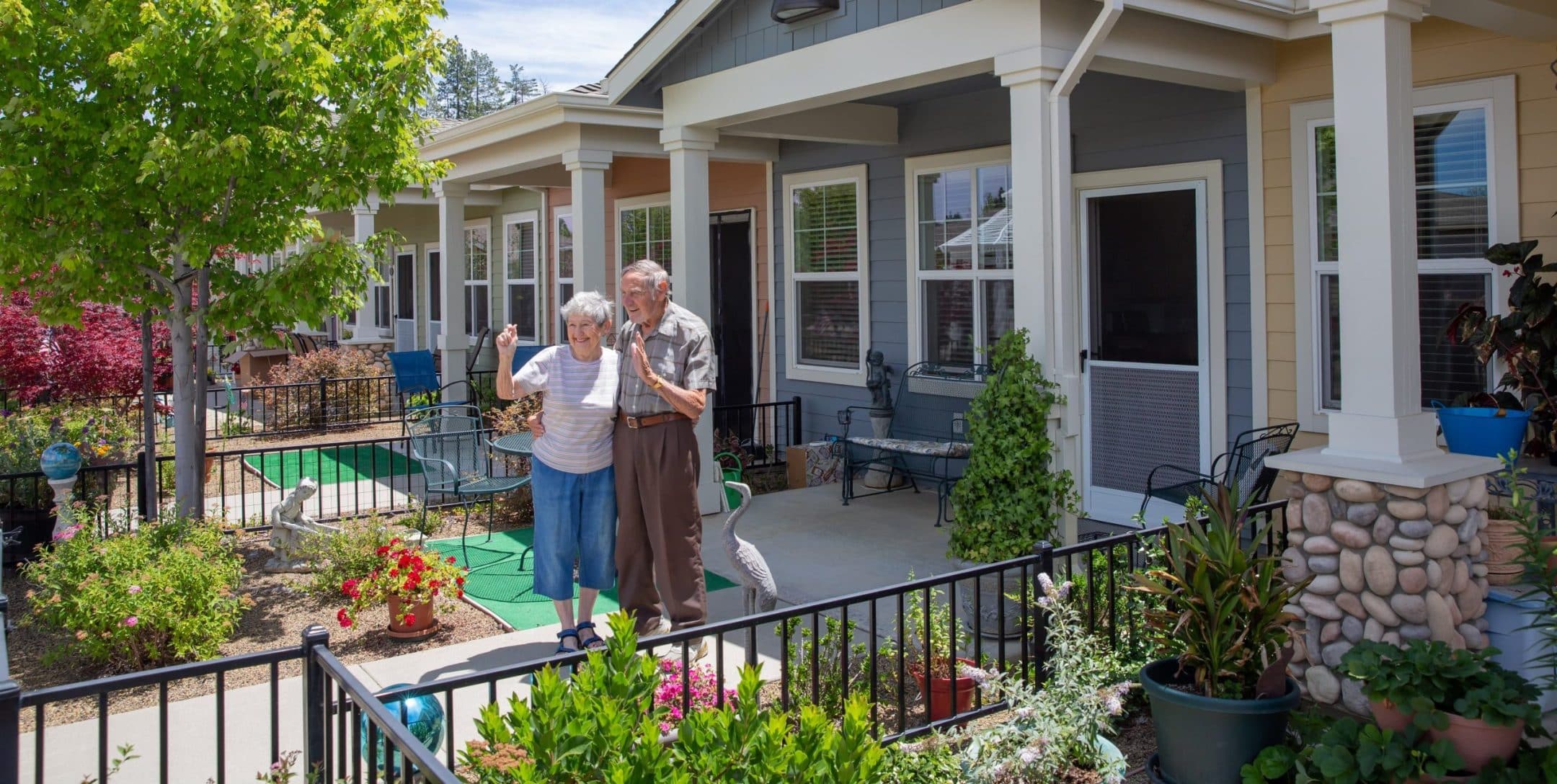 An elderly couple stands together and waves outside their porch in front of a row of colorful, single-story homes—showcasing inviting living options—surrounded by greenery and potted plants on a sunny day.