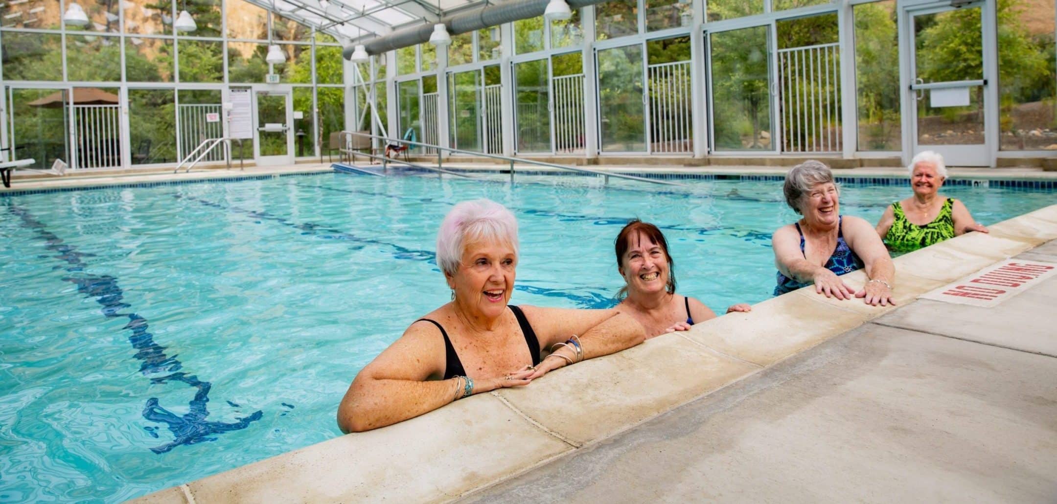 Four older women in swimsuits stand smiling at the edge of an indoor swimming pool with large glass windows, surrounded by greenery. The joyful, relaxed scene reflects resident culture built on friendship and active living.