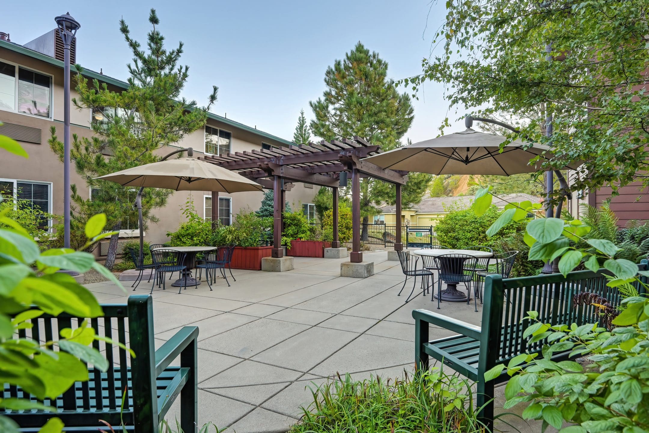 Outdoor patio area at Eskaton Village Placerville with green benches, round tables and chairs, large umbrellas for shade, lush plants and trees, and a wooden pergola in the center. Two residential buildings are in the background.