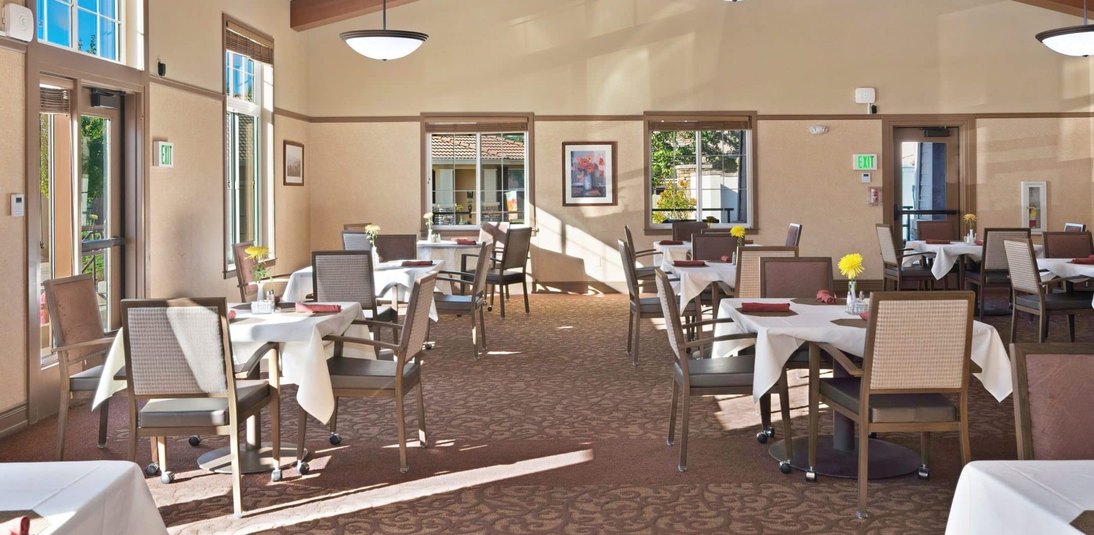 A spacious, well-lit Eskaton Village Placerville dining room features several tables with white tablecloths and yellow flowers in vases. Large windows fill the space with natural light, while wooden beams accent the ceiling. The room is empty of people.