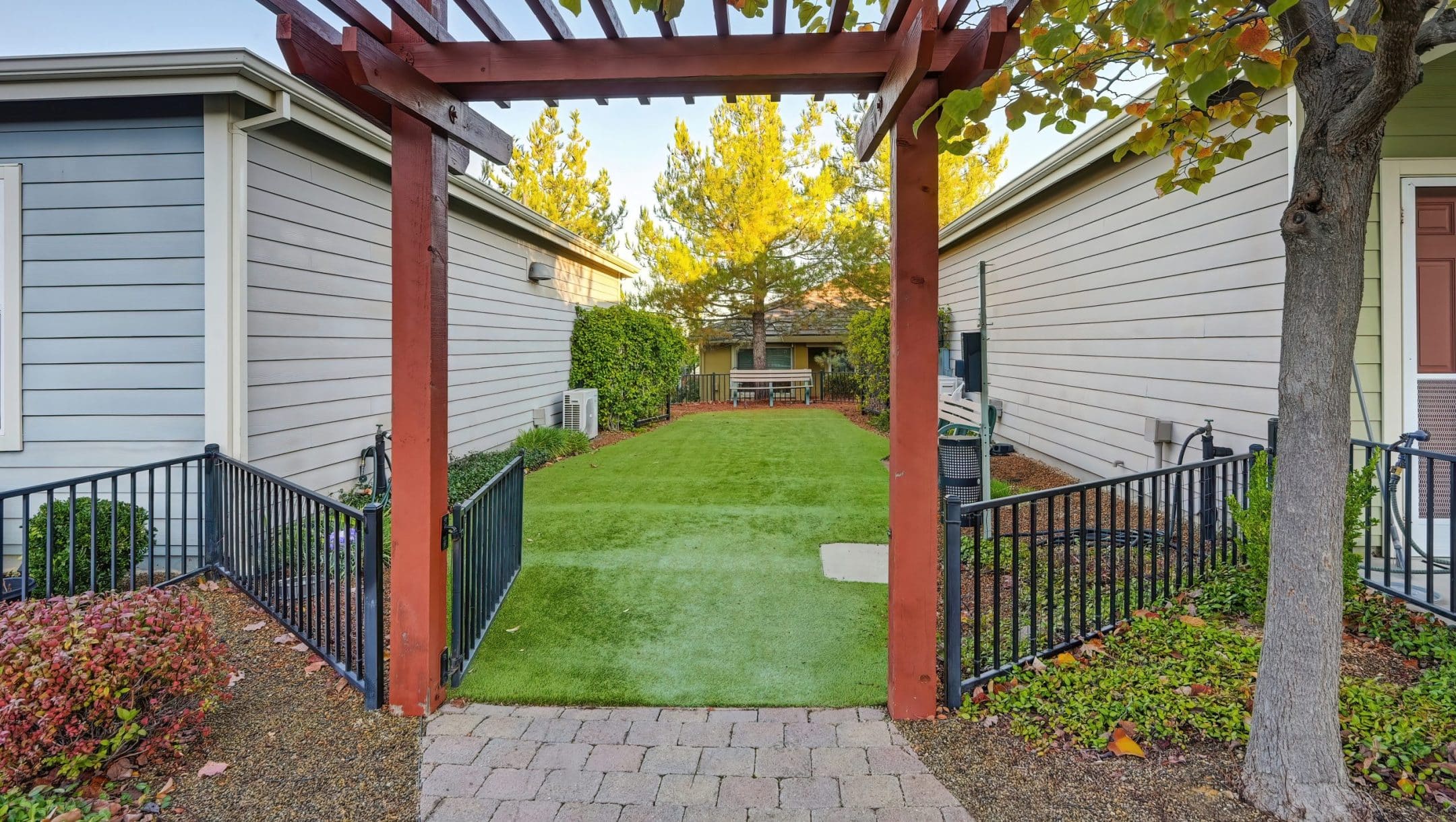 A wooden pergola stands over a brick pathway leading to a backyard with green artificial grass, highlighted by lush landscaping and two gray houses—one of the inviting Eskaton Village Placerville amenities under a clear sky.