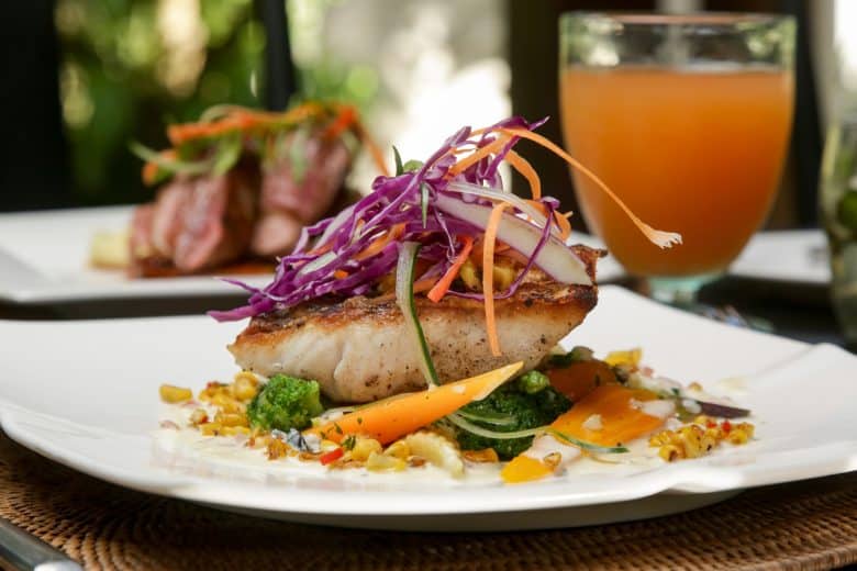A plated dish from Eskaton Village Placerville dining features grilled fish topped with shredded purple cabbage, carrots, and veggies, served over mixed sautéed vegetables, with a glass of orange beverage in the background.
