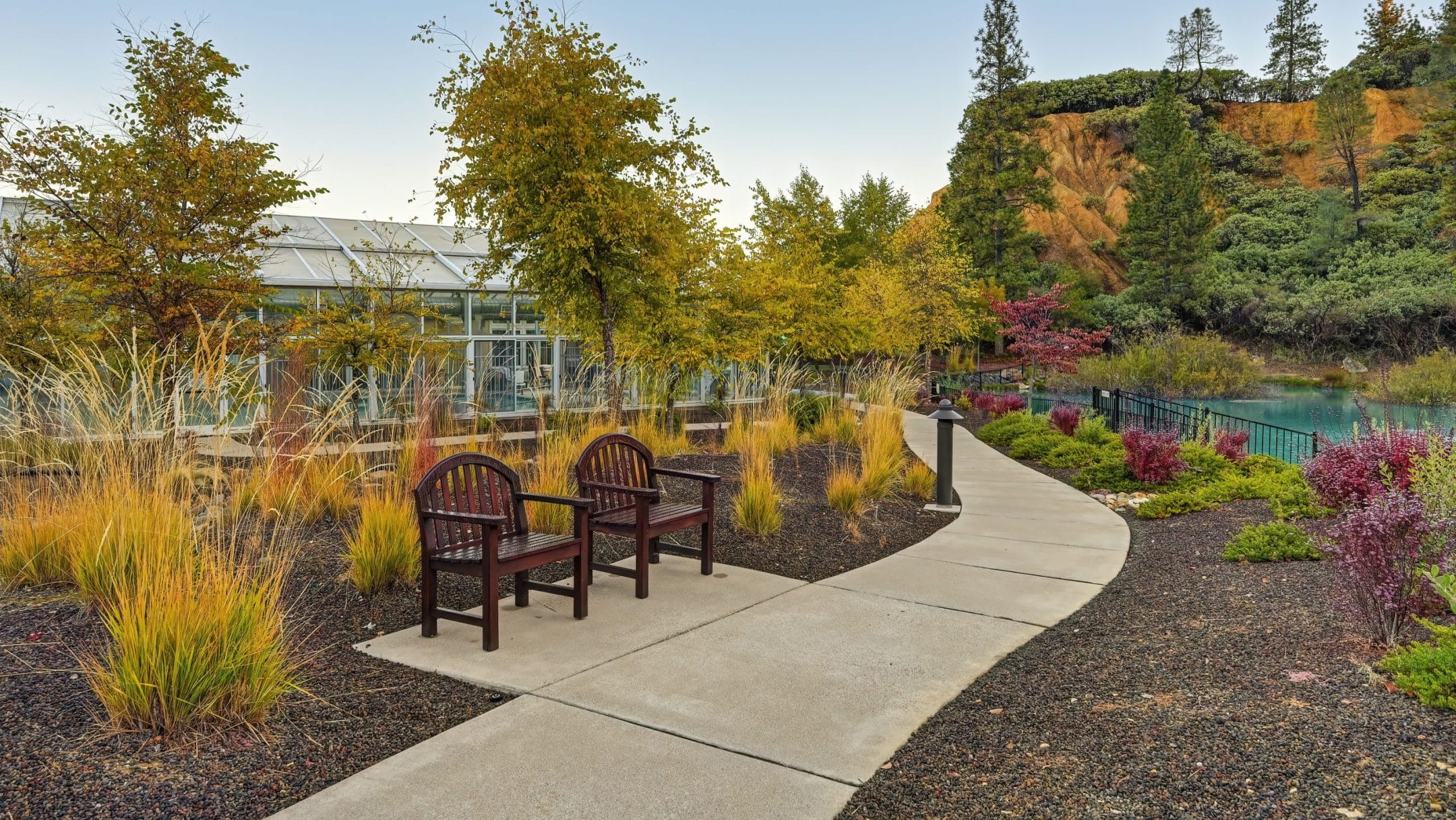 A curved concrete path with benches winds through a landscaped garden of autumn hues beside a glass greenhouse. This serene setting, part of Eskaton Village Placerville amenities, offers views of a rocky hill and evergreens in the background.