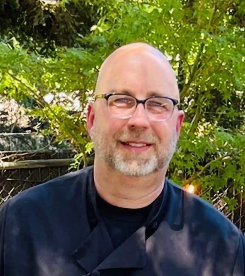 A smiling man wearing glasses and a black chef’s jacket that says “Phil Fiacco Chef” stands outdoors with trees, a fence, and the inviting atmosphere of Eskaton Village Placerville dining in the background.