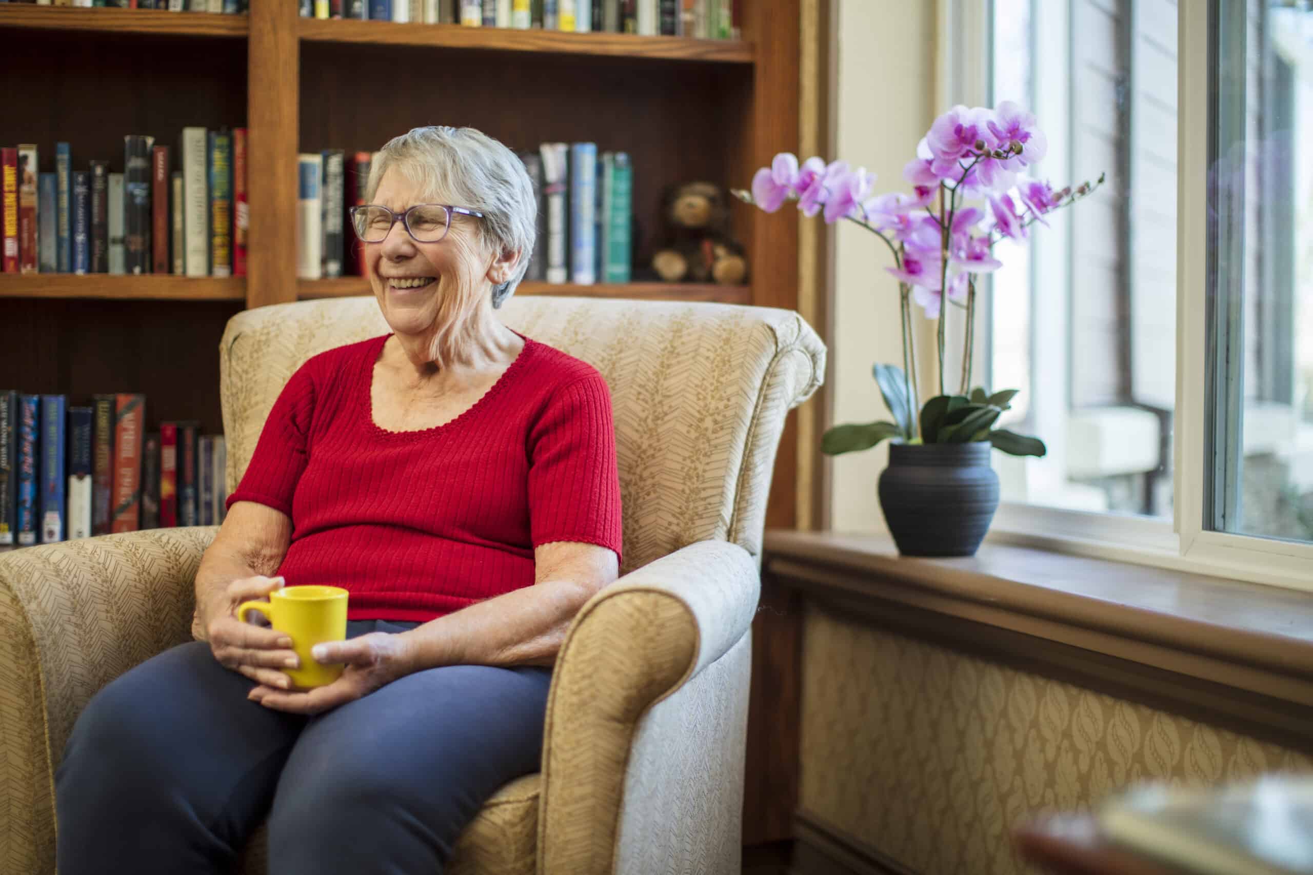 An older woman wearing glasses and a red shirt sits smiling in an armchair, holding a yellow mug. Behind her is a bookshelf, and a pink orchid sits by a window—a warm scene from our Community Spotlight Event.