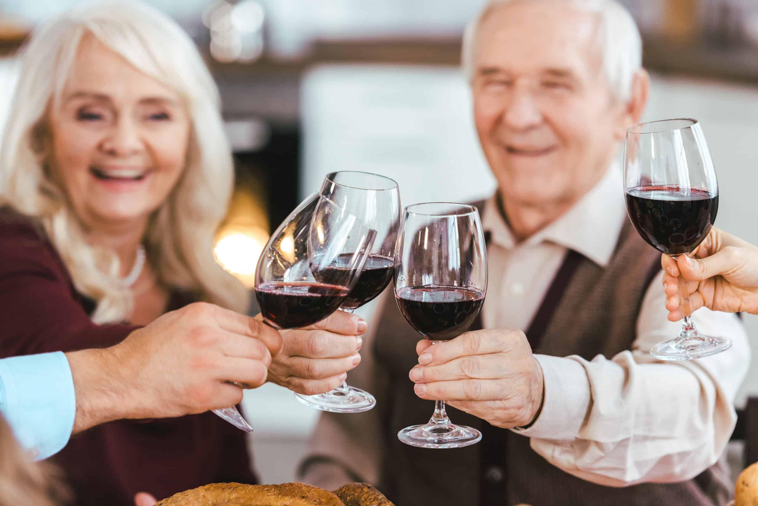 Two smiling older adults and others out of frame cheerfully clink glasses of red wine together, celebrating around a table in a cozy indoor setting during a Community Spotlight Event.