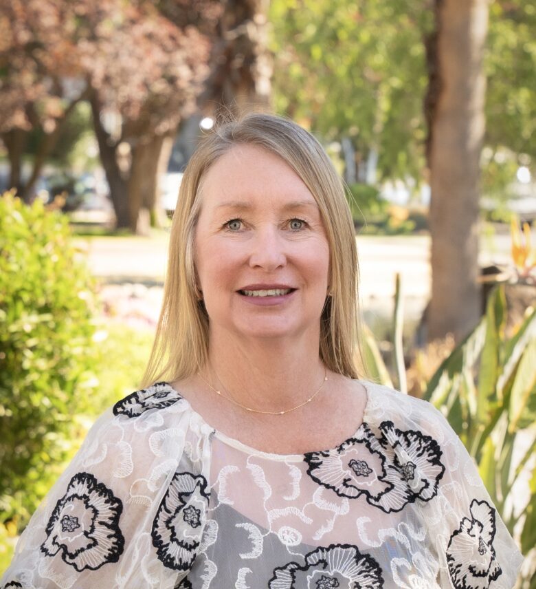 A woman with straight blonde hair and a floral-patterned sheer blouse smiles while standing outdoors in a sunlit garden at Eskaton Village Placerville, surrounded by trees and greenery in the background.