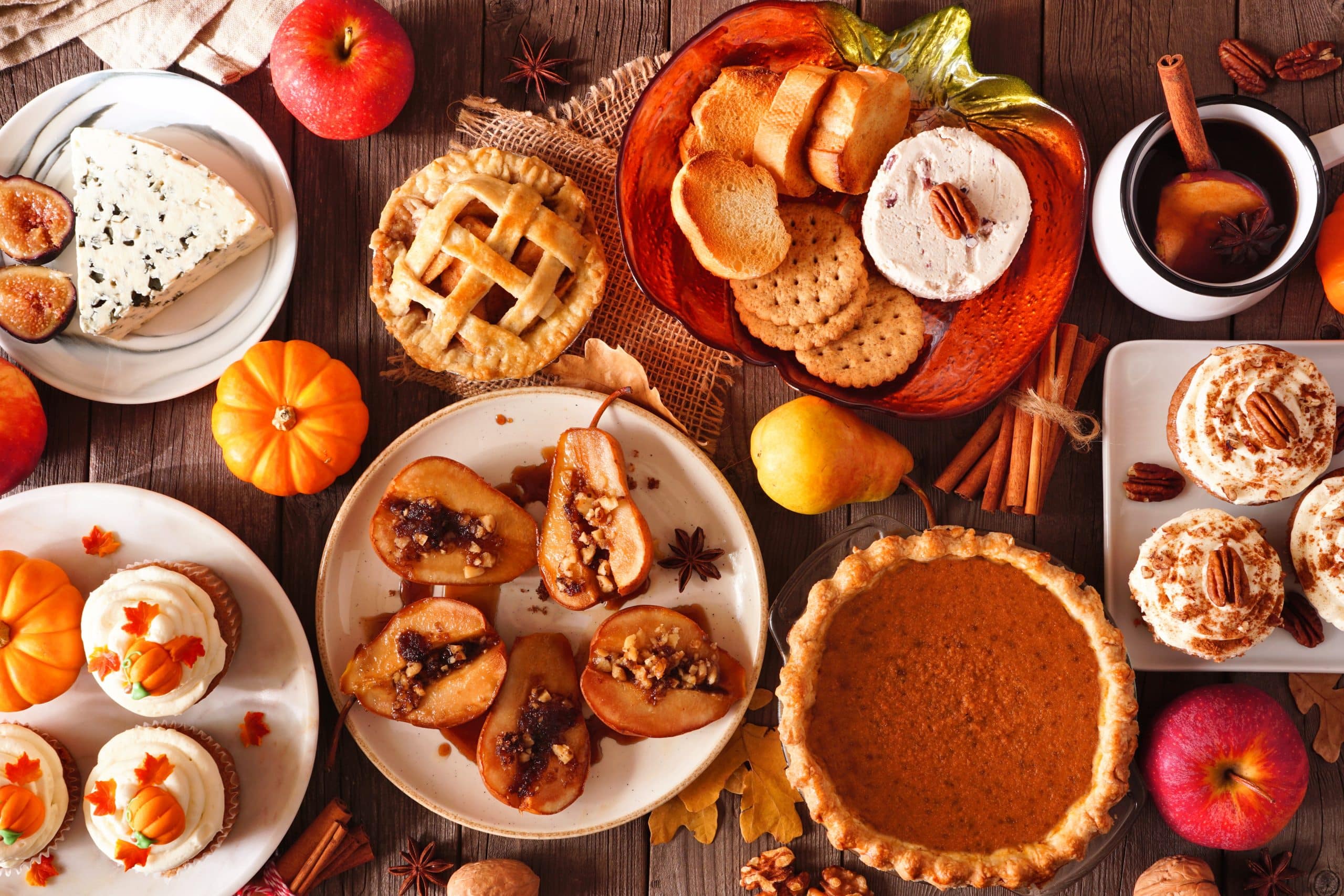 An overhead view of a rustic autumn table set for an Autumn Pie Paint Social, featuring pumpkin pie, baked pears, cheese, apple pie, cupcakes, crackers, apples, and small pumpkins—all beautifully decorated with autumn leaves and spices on a wooden surface.
