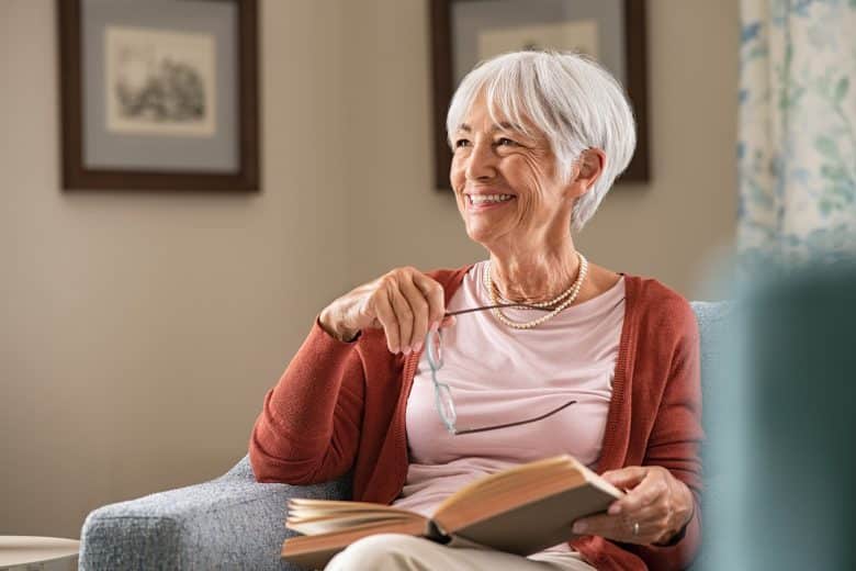 Smiling older woman with short white hair sits on a blue armchair, holding eyeglasses and an open book, wearing a coral cardigan and pearl necklace in a cozy, softly lit room that reflects the warm and inviting Eskaton Village Placerville lifestyle.