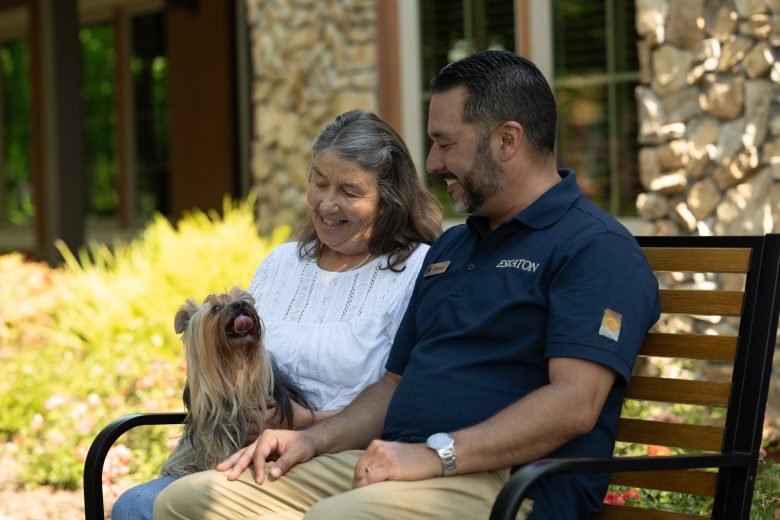 An older woman and a man sit on a bench outside an assisted living community, smiling and petting a small, fluffy dog with its tongue out. They appear relaxed and happy, surrounded by greenery and a stone building.