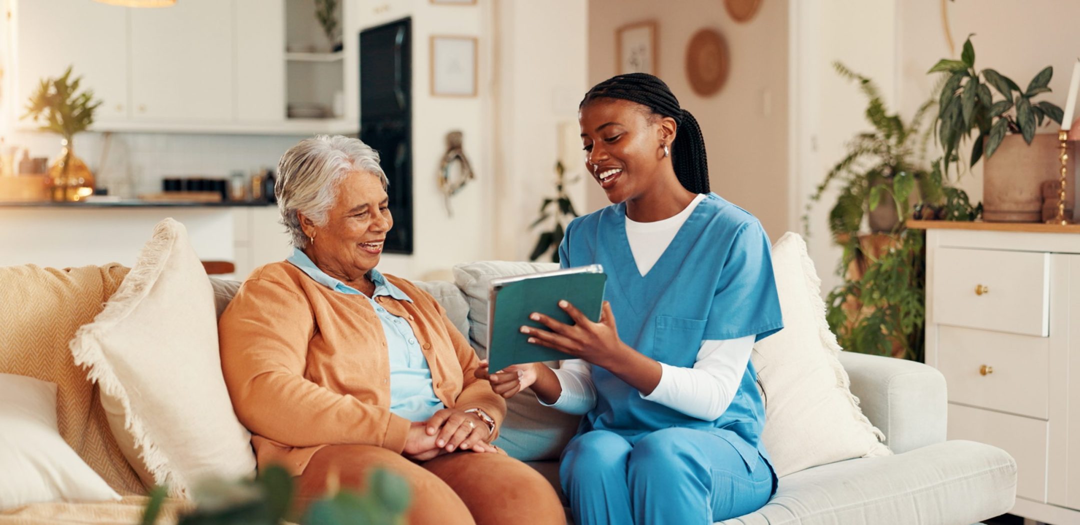 A nurse in blue scrubs sits on a couch with an elderly woman, smiling as they look at a book together in a cozy, well-lit living room with plants and neutral decor.
