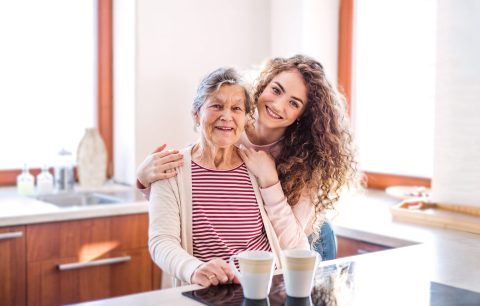 A smiling elderly woman and a young woman with curly hair embrace in a bright kitchen, standing behind a counter with two mugs. Sunlight streams in through large windows, creating a welcoming homepage feel.