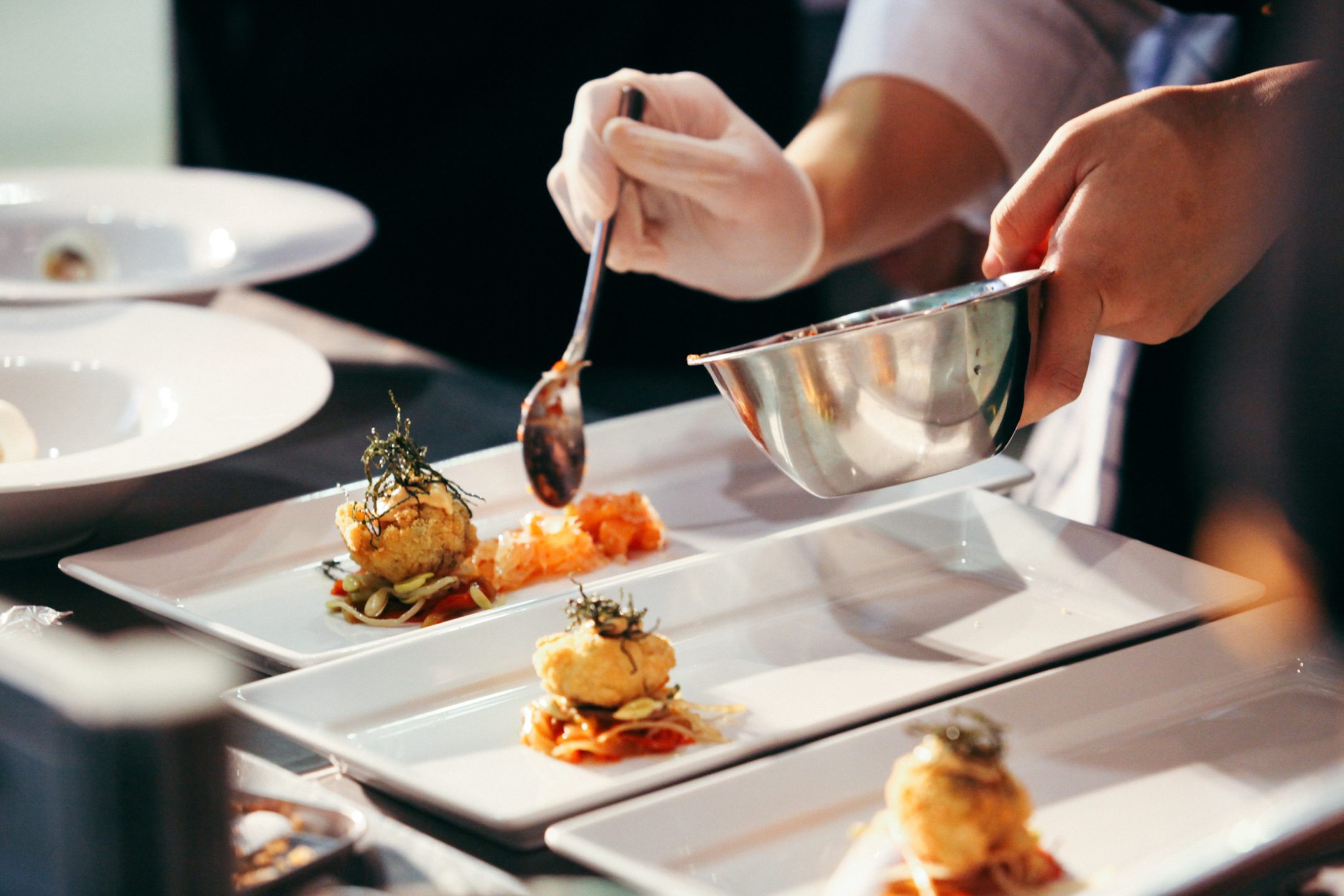 A chef wearing gloves uses a spoon to plate food, adding a garnish or sauce to elegantly arranged appetizers on rectangular white plates, creating an exceptional culinary experience in a professional kitchen.