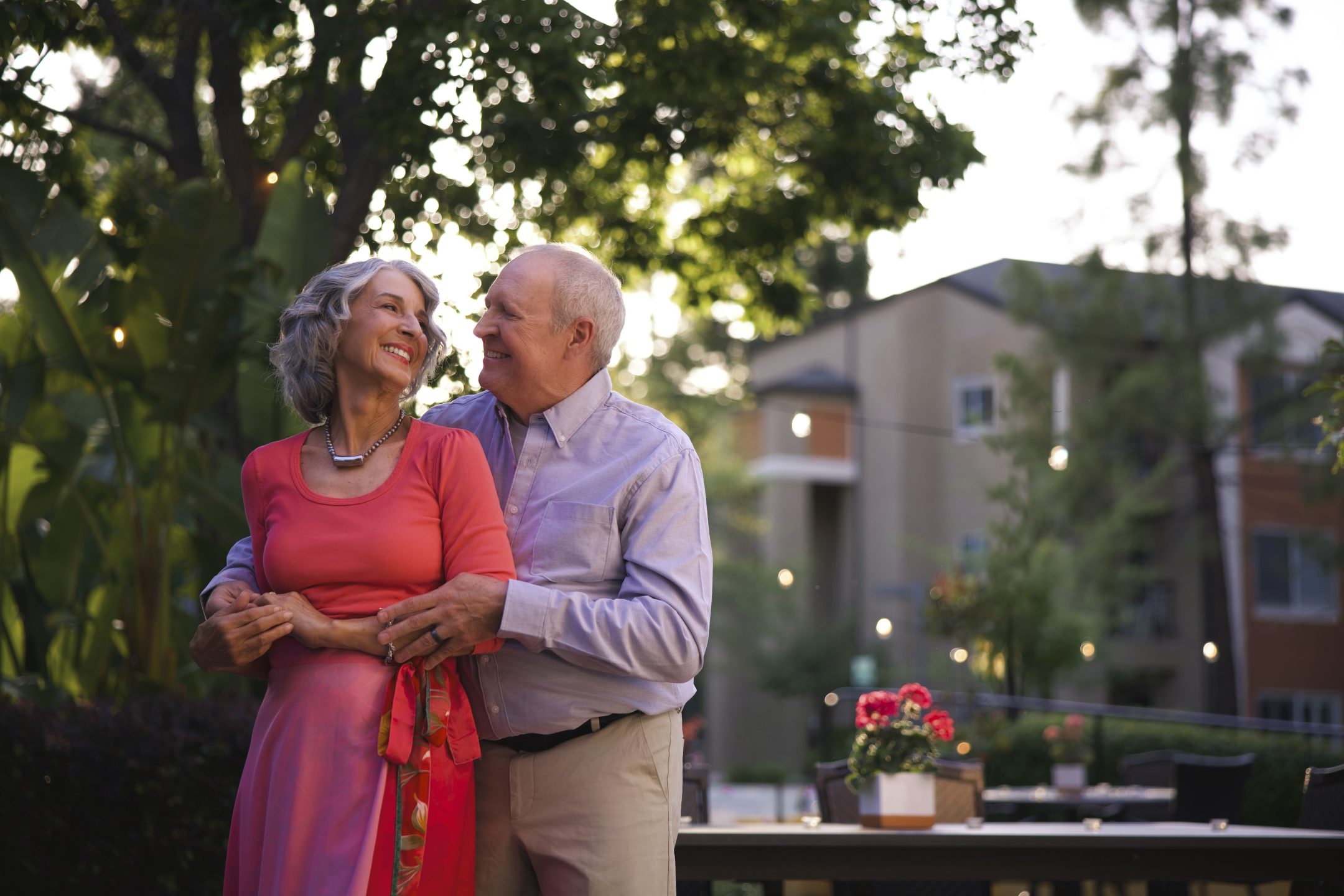 An older couple stands outside, smiling and embracing, with greenery and a modern apartment building in the background. The woman wears a pink dress and the man wears a light shirt and pants. Evening light creates a warm, cheerful atmosphere.