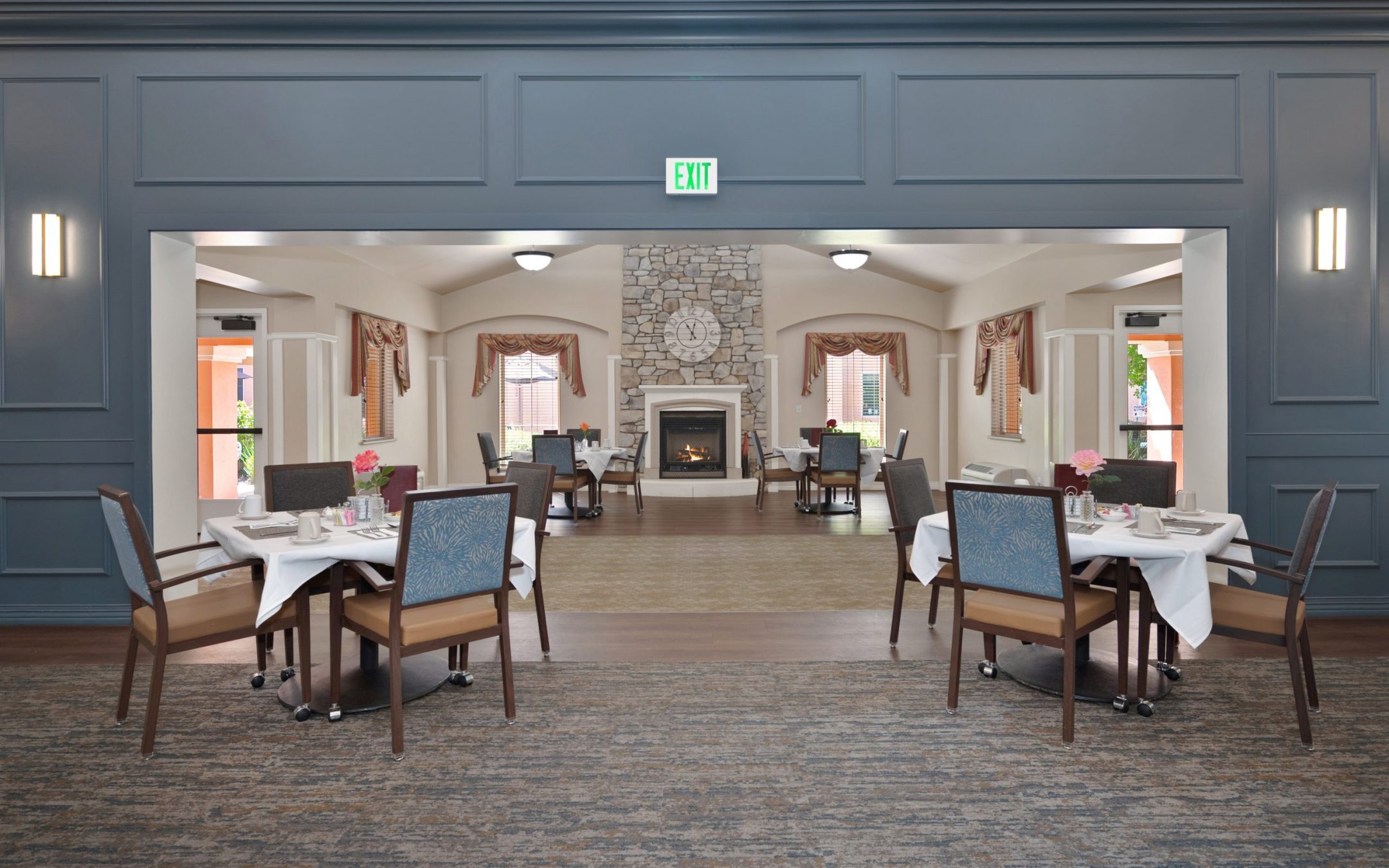 A spacious dining area with neatly set tables and chairs, leading to a lounge with a stone fireplace, clock, and large windows with drapes, all under a high ceiling and soft lighting.