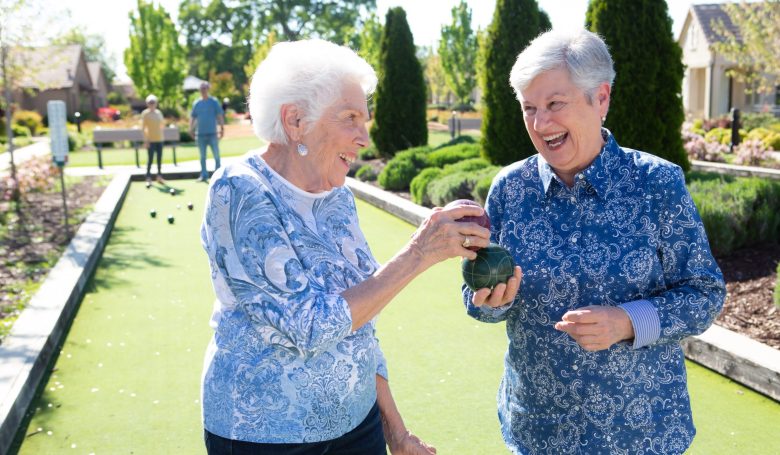 Two older women smiling and holding a bocce ball on an outdoor bocce court, with other people playing in the background on a sunny day.