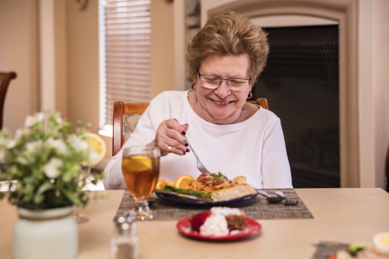 An older woman wearing glasses and a white top smiles as she eats a meal at a dining table, with a plate of food, iced tea, and a floral centerpiece in front of her.