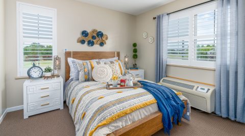 A cozy, brightly lit bedroom features a neatly made bed with striped yellow and gray bedding, decorative pillows, a blue throw, two white nightstands, windows with shutters, and cheerful wall art above the headboard.