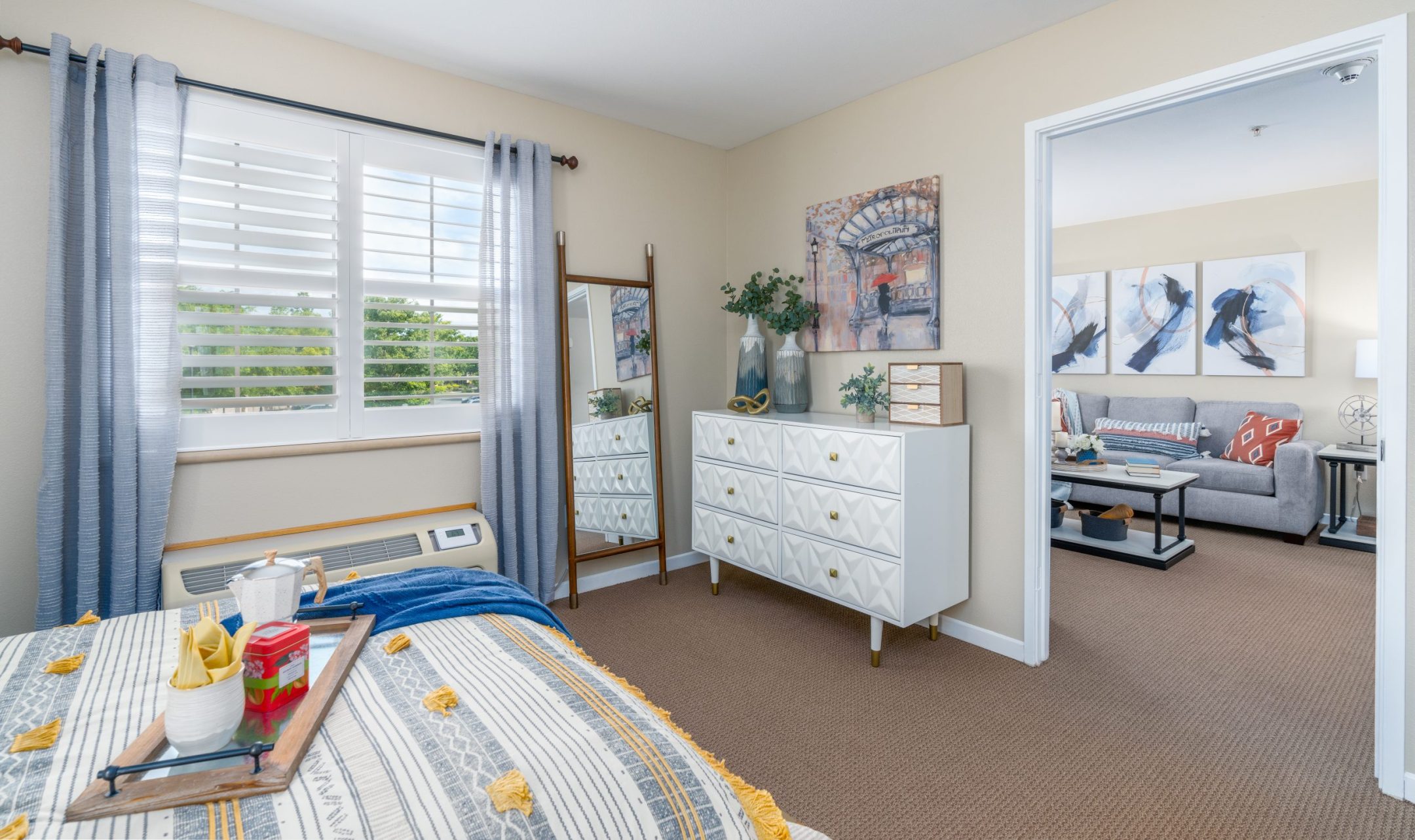 A bright bedroom with beige walls, a bed with a striped blanket, a white dresser with decor and plants, a tall mirror, and a window with sheer curtains. An adjacent living room is visible through an open door.