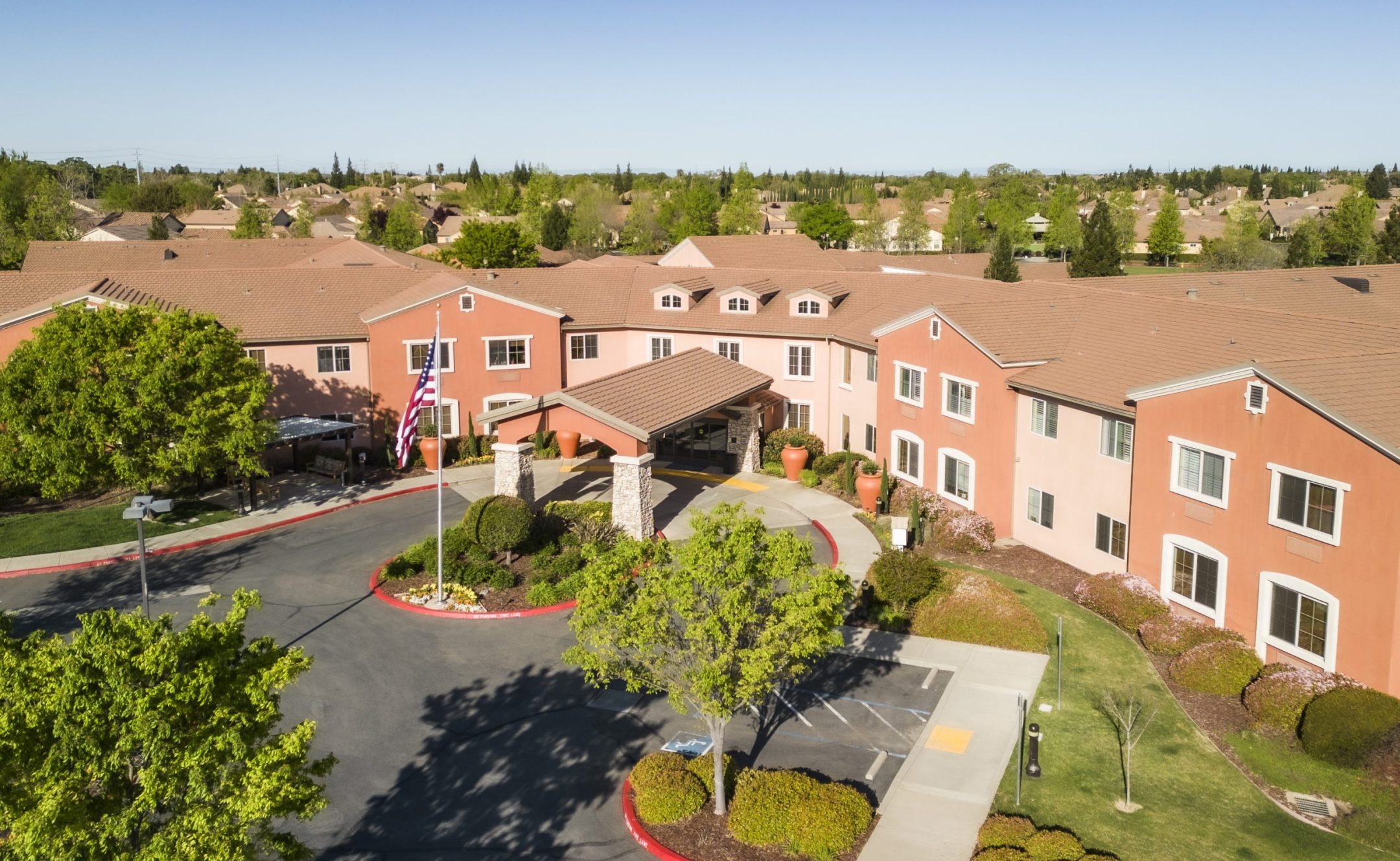 Aerial view of a large, two-story, terracotta-colored building with a covered entrance, surrounded by trees, manicured lawns, and a parking lot. An American flag is displayed near the entrance.