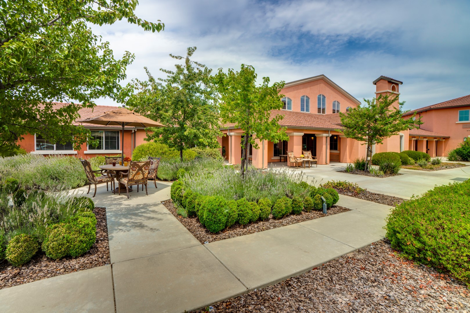 A landscaped courtyard with trimmed bushes and trees, surrounded by orange stucco buildings. Outdoor seating areas with tables and chairs are placed on walkways under partly cloudy skies.