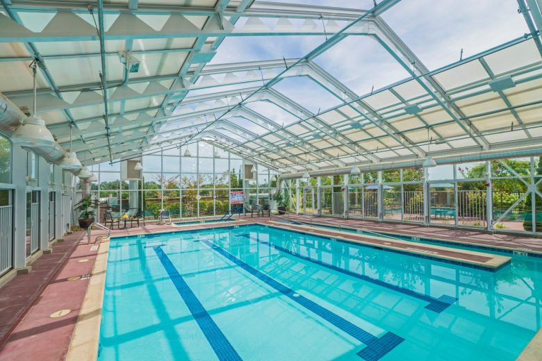An indoor swimming pool with clear blue water under a large glass ceiling and walls, allowing natural light to fill the space. Poolside chairs and greenery are visible outside the glass enclosure.