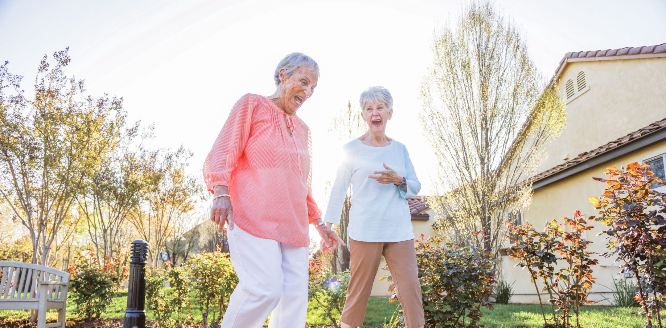 Two older women are laughing and walking together on a sidewalk in a sunny, landscaped neighborhood with trees and bushes around them. One wears a pink top and white pants; the other wears a light blue top and tan pants.