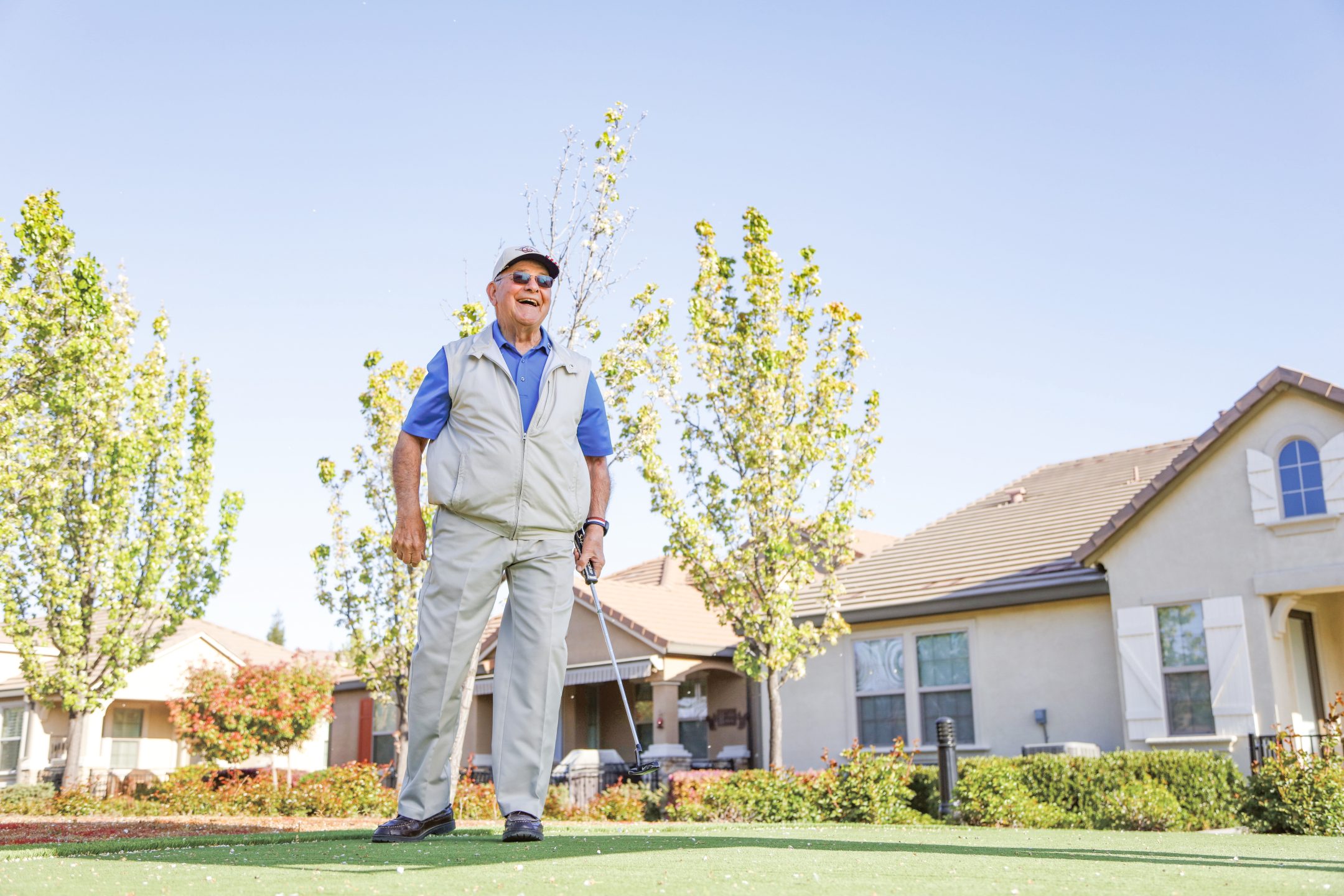 An older man wearing sunglasses and a vest smiles while standing on a putting green with a golf club, with houses and trees in the background on a sunny day.
