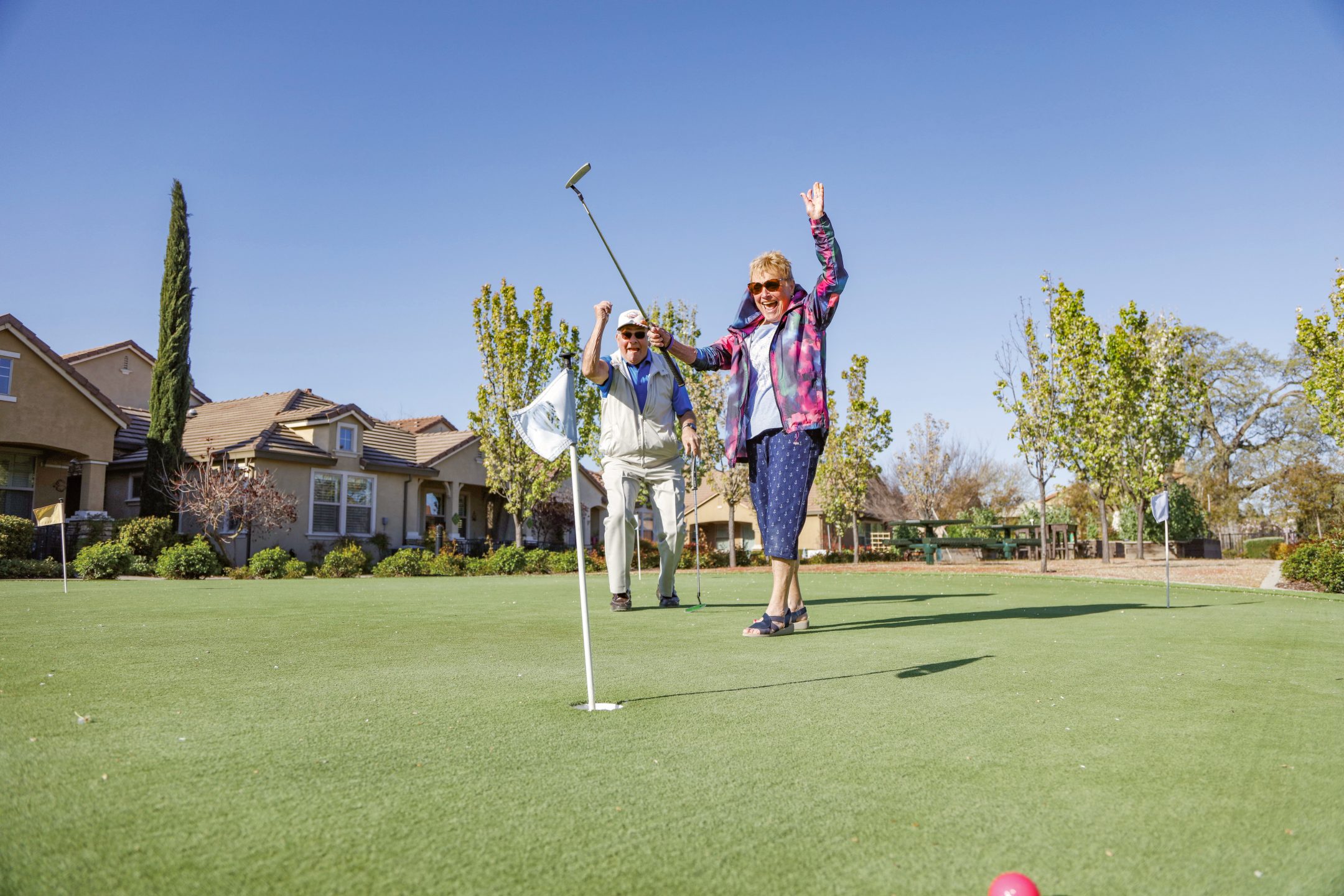 Two older adults cheer and celebrate near a golf hole on a sunny putting green, surrounded by houses and trees, with one person holding a golf club and the other making a peace sign.