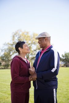 An older couple wearing athletic outfits smiles and holds hands while standing outside on a grassy field, with trees and a clear sky in the background.