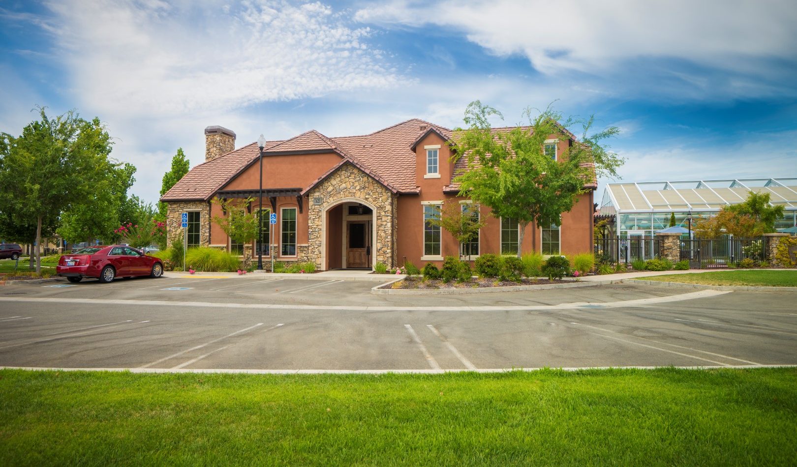 A large, tan house with stone accents and a red-tiled roof sits beside a greenhouse and a parked red car, surrounded by trees, greenery, and a wide driveway under a partly cloudy sky.