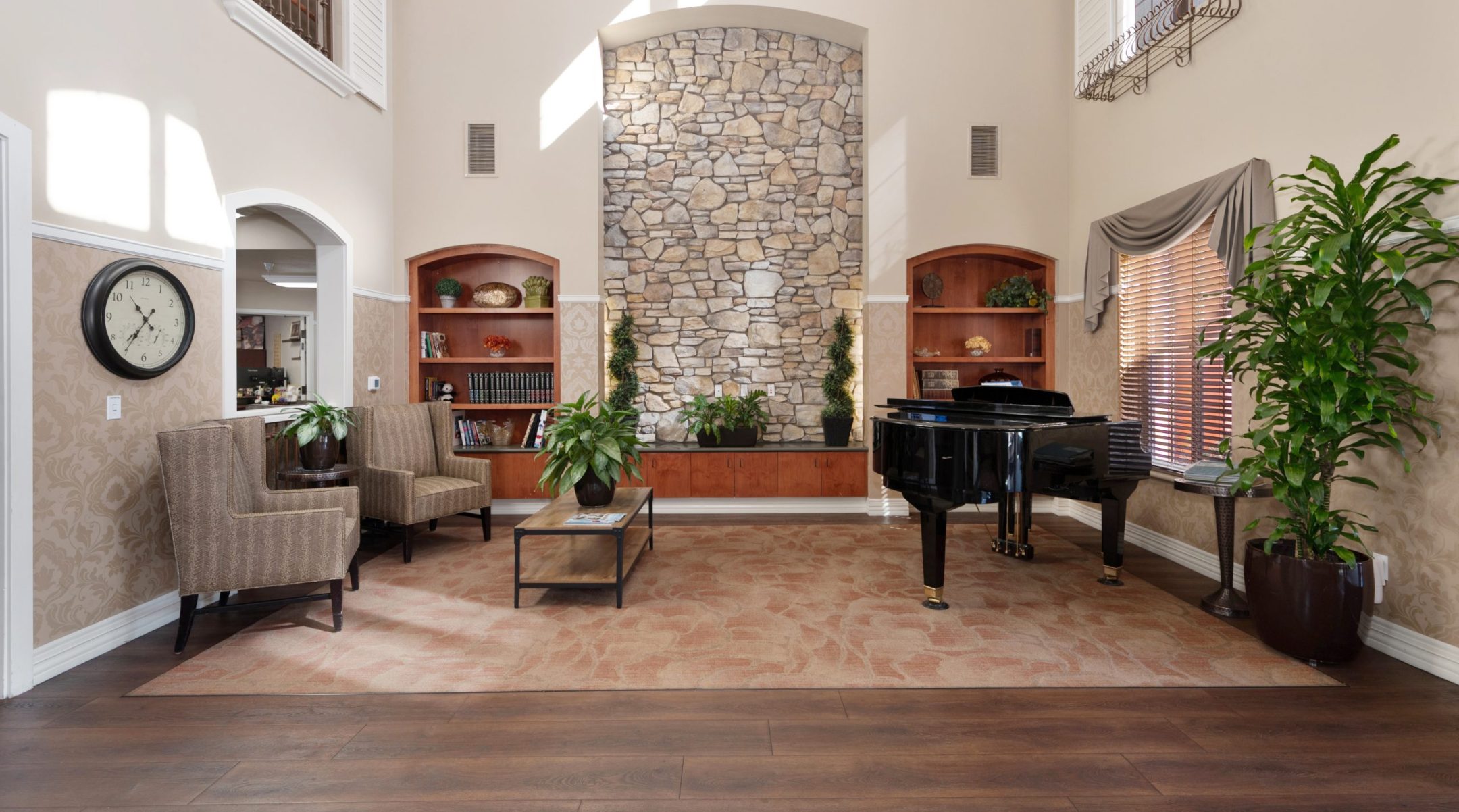 A spacious living room with high ceilings, a stone accent wall, built-in shelves, a grand piano, armchairs, potted plants, a wall clock, and large windows with blinds allowing sunlight inside.