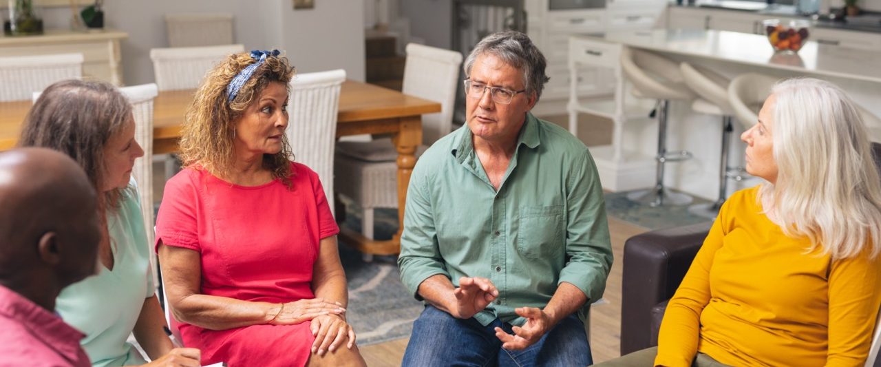 A group of five adults sit in a circle in a bright room at Eskaton Village Placerville memory care, engaged in a serious conversation. One man in a green shirt gestures while speaking as the others listen attentively.