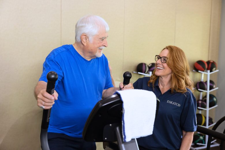 An Eskaton Village Roseville resident enjoys exercising on a stationary machine, smiling and chatting with a woman in glasses and an ESKATON shirt. A rack of medicine balls is visible in the background, highlighting the vibrant resident experience.
