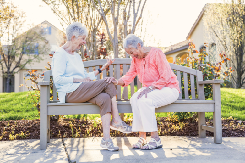 Two older women with gray hair sit on a wooden bench outside Eskaton Village Roseville senior living, smiling and laughing together in the sunshine. One touches the other’s knee warmly, with trees, grass, and houses in the background.
