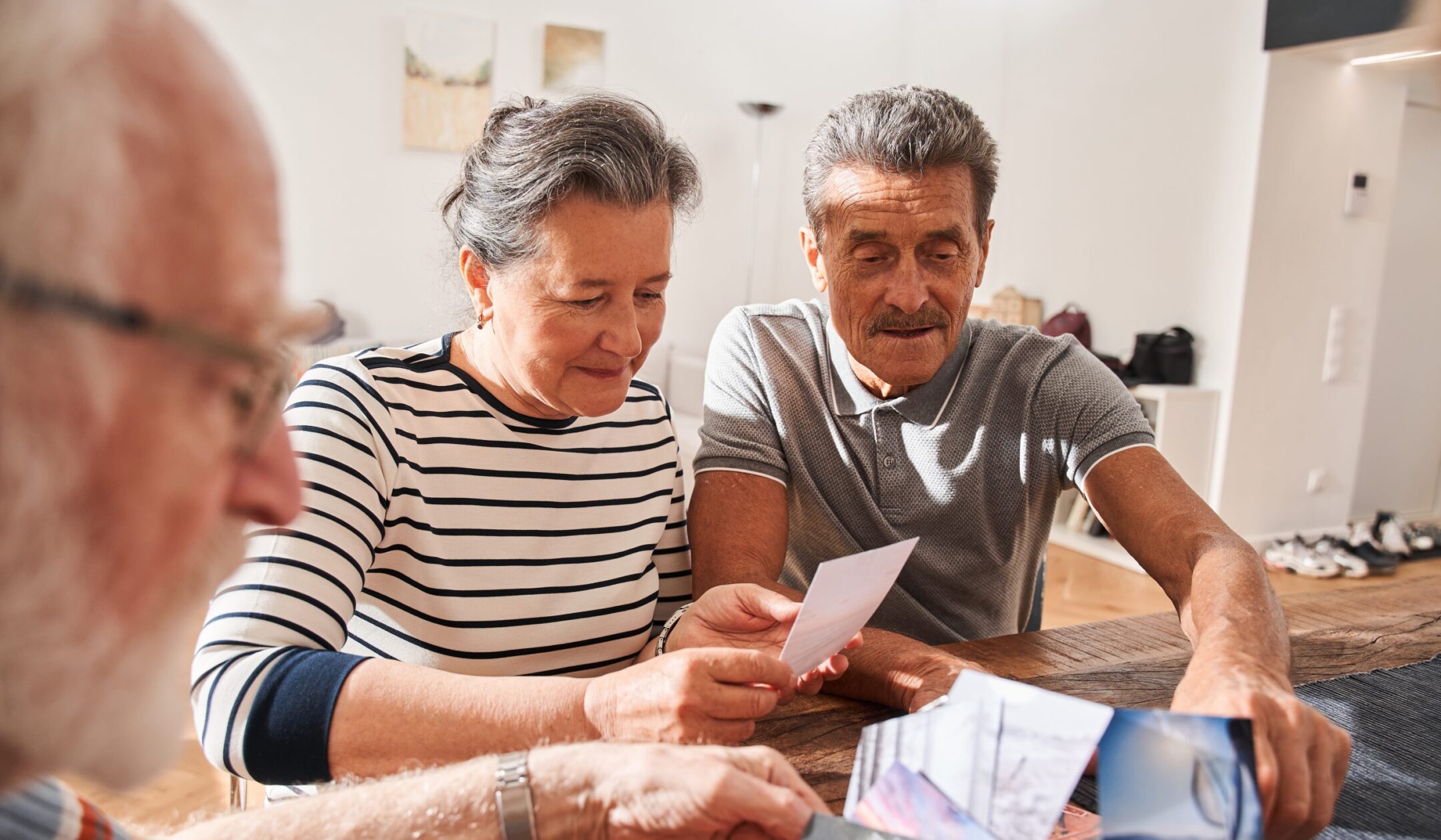 Three older adults sit at a table indoors, smiling and looking at photographs together. Sunlight streams into the room, creating a warm and cozy atmosphere. Memory cafe.