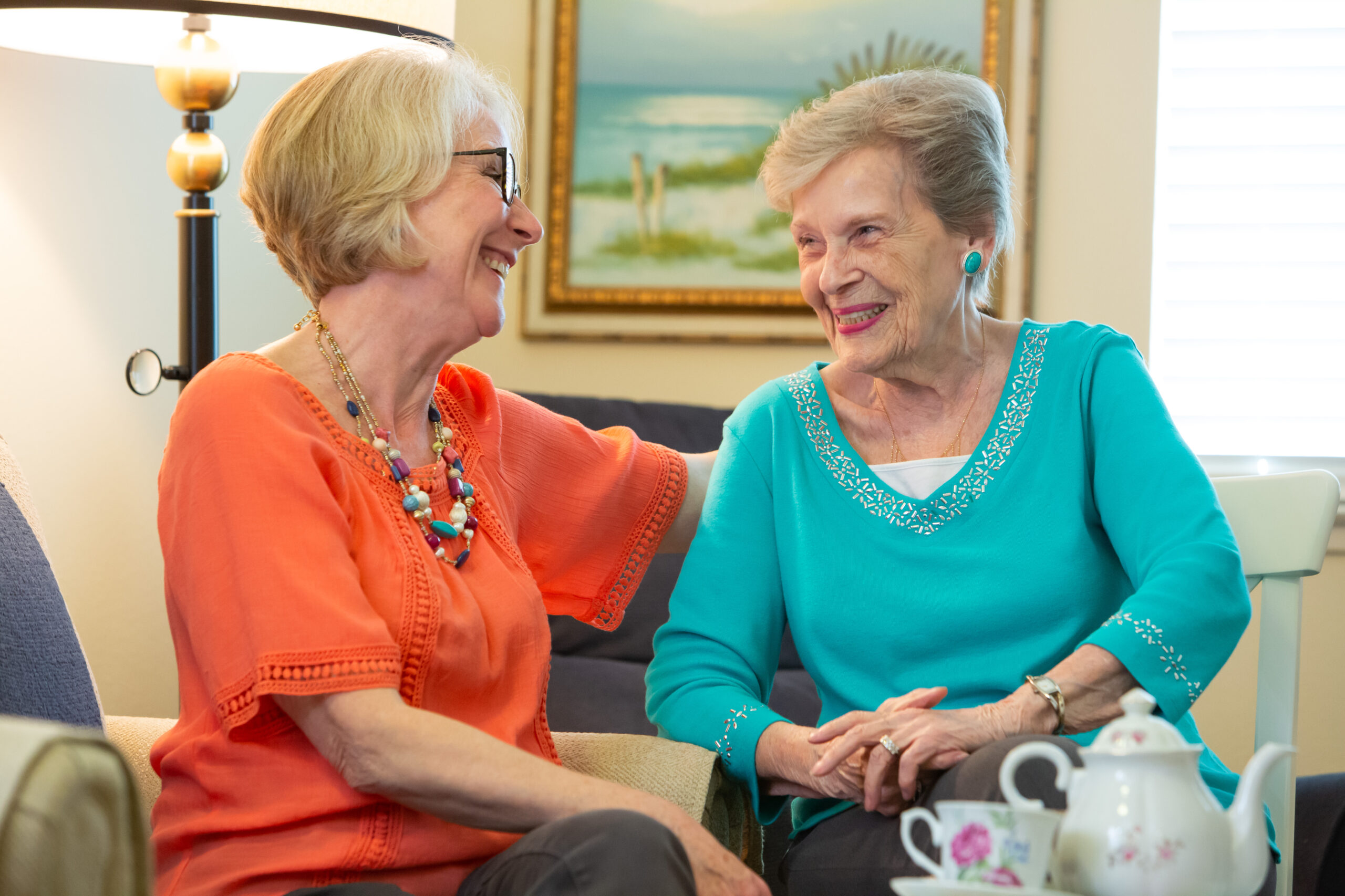 Two elderly women sit on a couch at Enrichment Cafe, smiling and chatting together. One wears an orange top and glasses, the other a turquoise top. A tea set is on the table, with a painting hanging on the wall behind them.