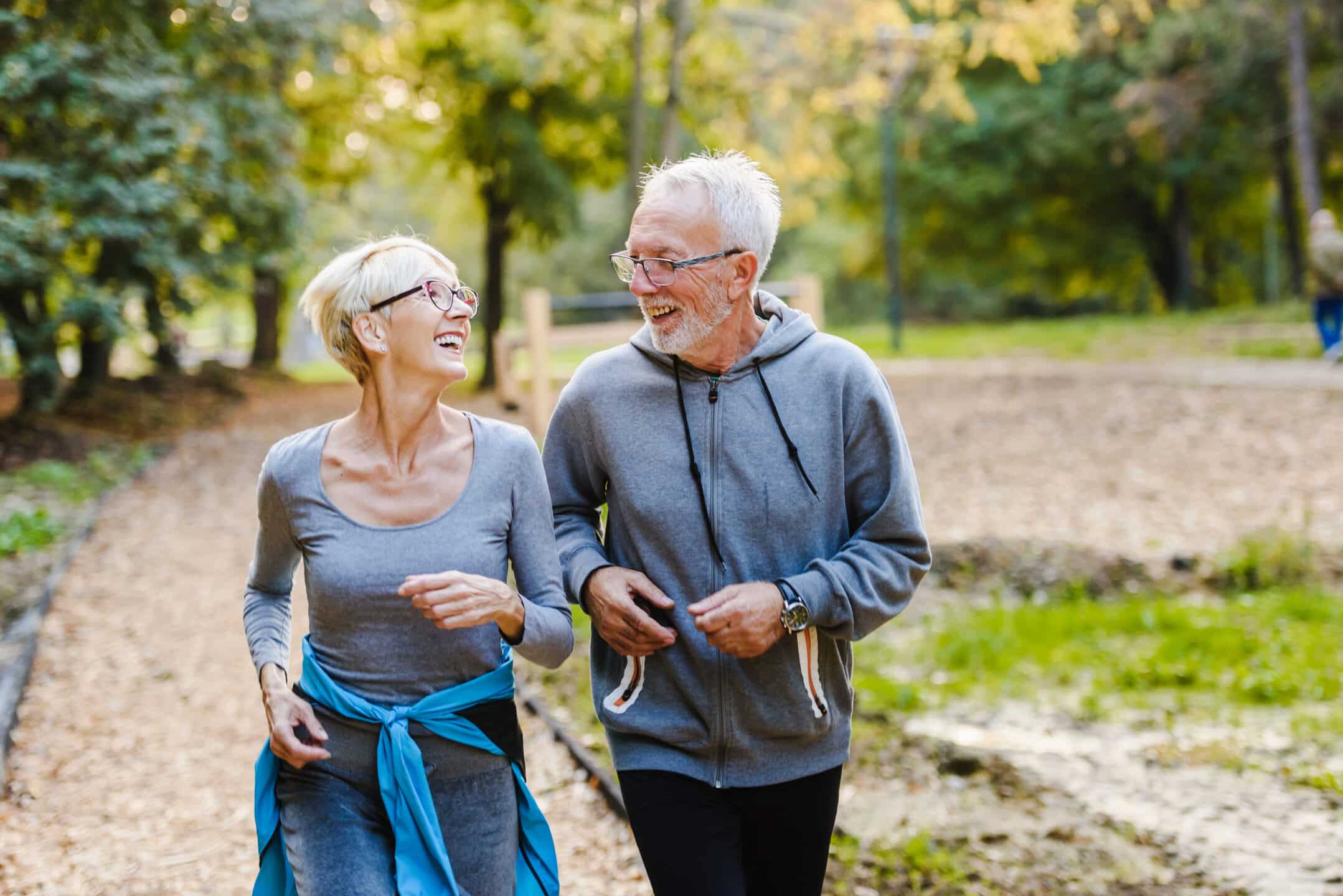 An older couple in athletic wear smiles and walks together on a wooded park path, enjoying a sunny day after attending the Heath & Wellness fair. The woman has short blonde hair and glasses, and the man has gray hair and a beard.