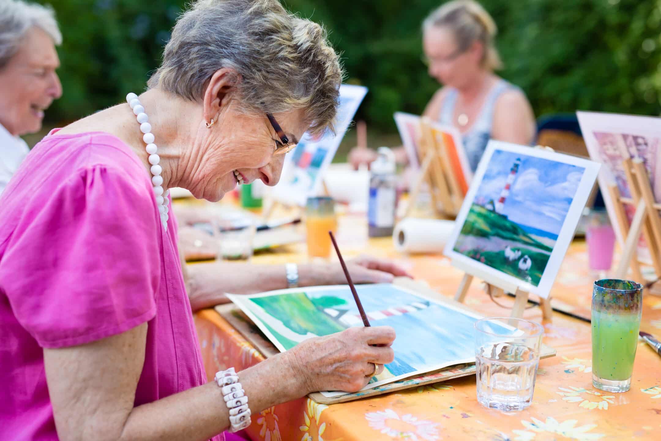 Elderly women painting outdoors at a Paint & Sip event, smiling and focused on their colorful landscape artwork, with easels, art supplies, and drinks nearby on a sunny day.