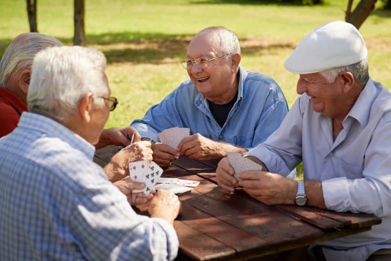 Four elderly men sit outdoors at a wooden table, laughing and playing cards together on a sunny day, surrounded by green grass and trees—a wonderful reflection of the vibrant Eskaton Village Roseville lifestyle.