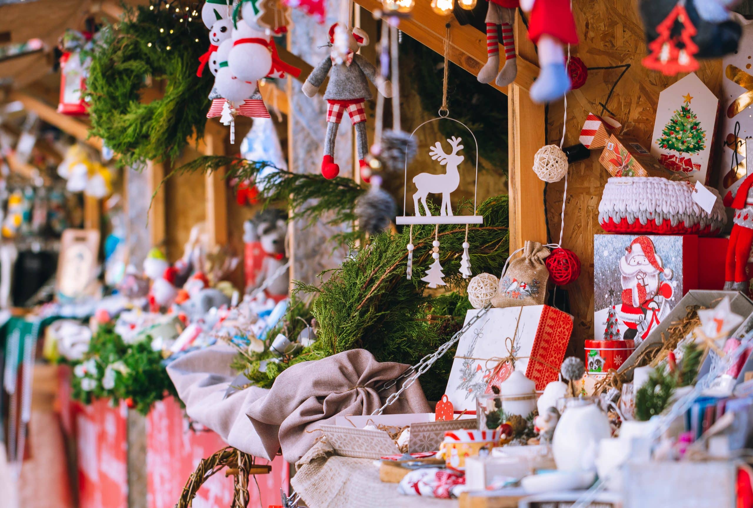 A festive Christmas market stall at the annual holiday craft fair displays ornaments, greenery, plush toys, and holiday decorations, with a focus on red and white colors and a wooden reindeer hanging in the center.